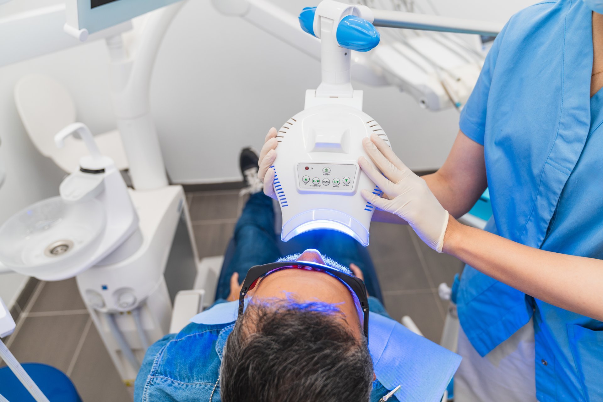 A man is getting his teeth whitened in a dentist's office. A woman is assisting him.