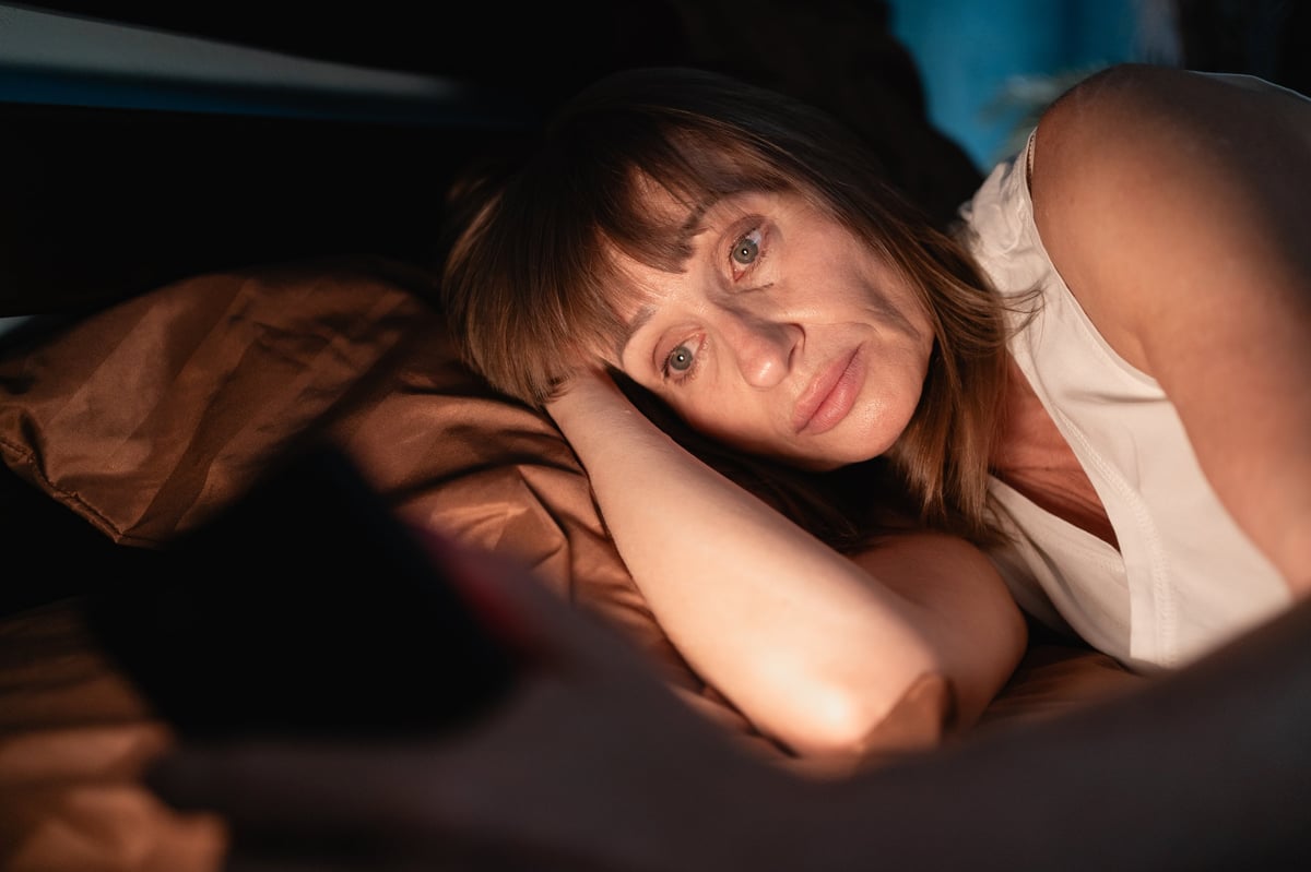 Sleepy woman awake at night using smart phone lying in bed in her bedroom. Using mobile for chatting. Copy space