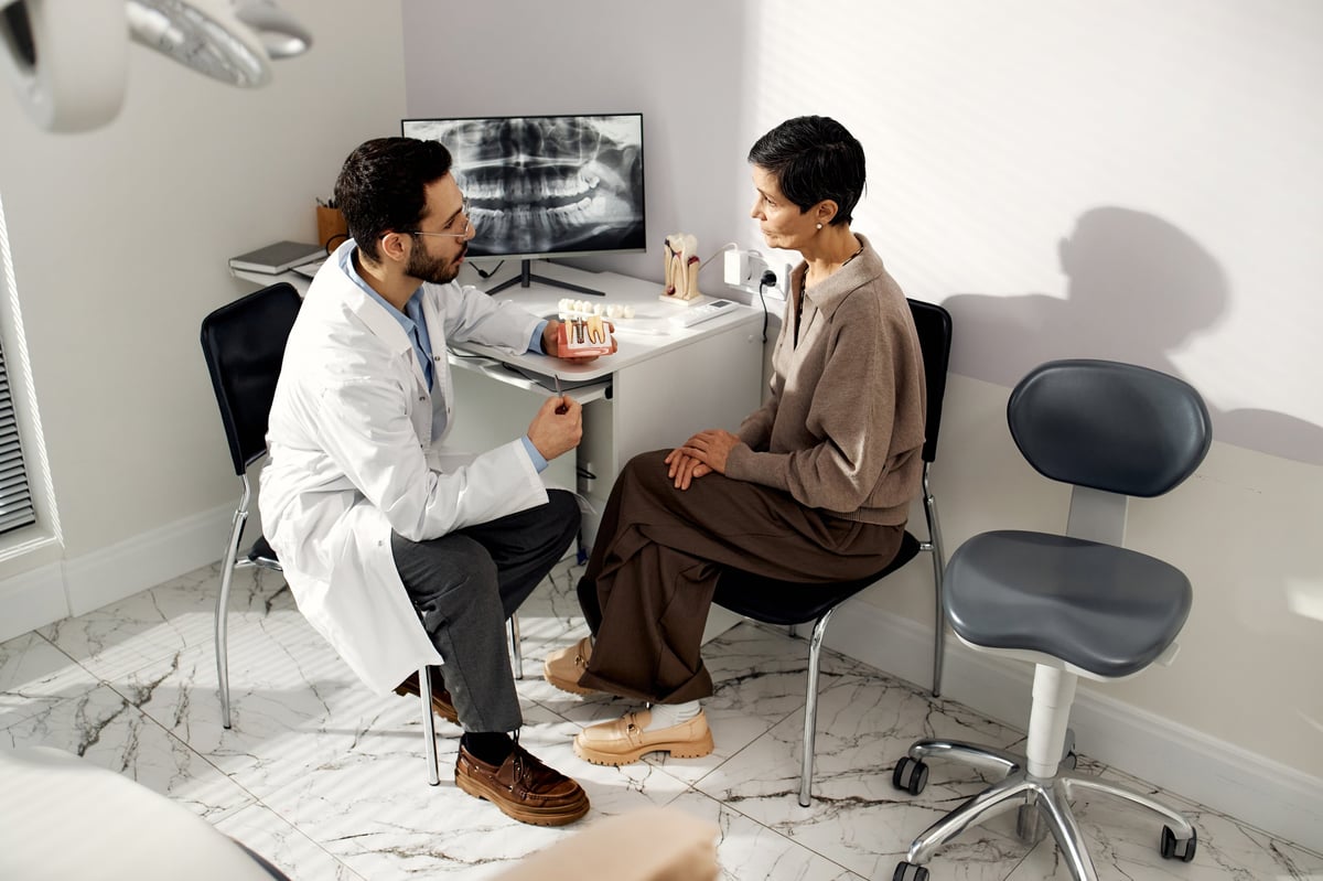 Middle Eastern dentist explaining dental procedure to middle aged Caucasian patient in dental office, both sitting and discussing treatment plan with dental x-ray visible in background