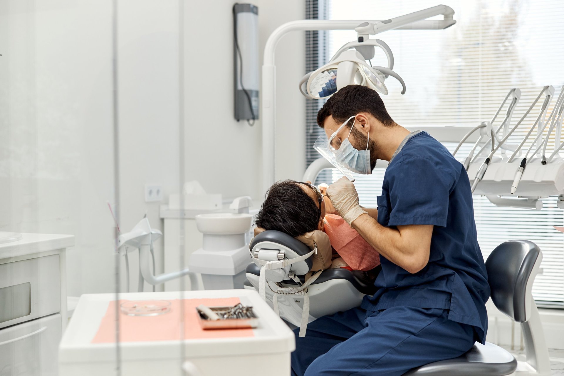 Middle Eastern male dentist examining patient in dental chair during routine checkup in modern dental office, both wearing protective masks, dental tools visible in foreground.