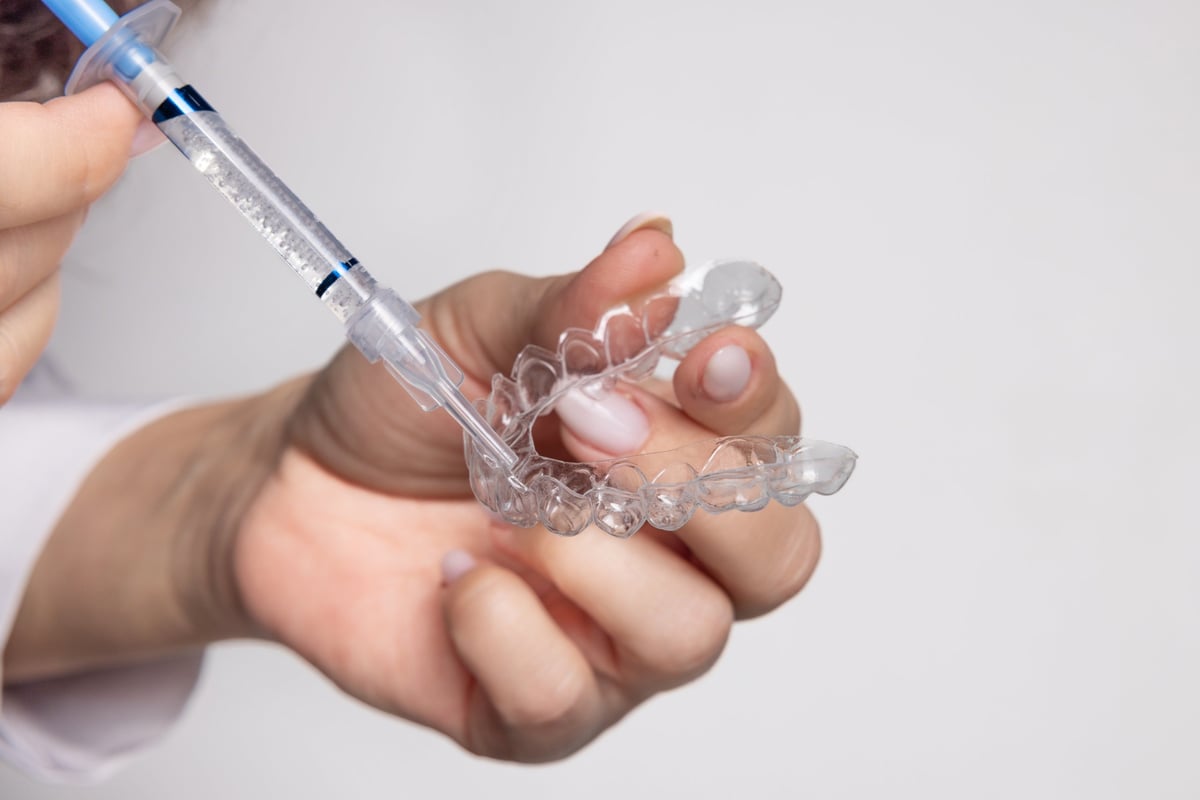 Woman holds whitening trays and solution on a minimal backdrop. Clear trays, dental model, case and syringes placed on the table. The woman applies gel into the trays for tooth care and bright smile.