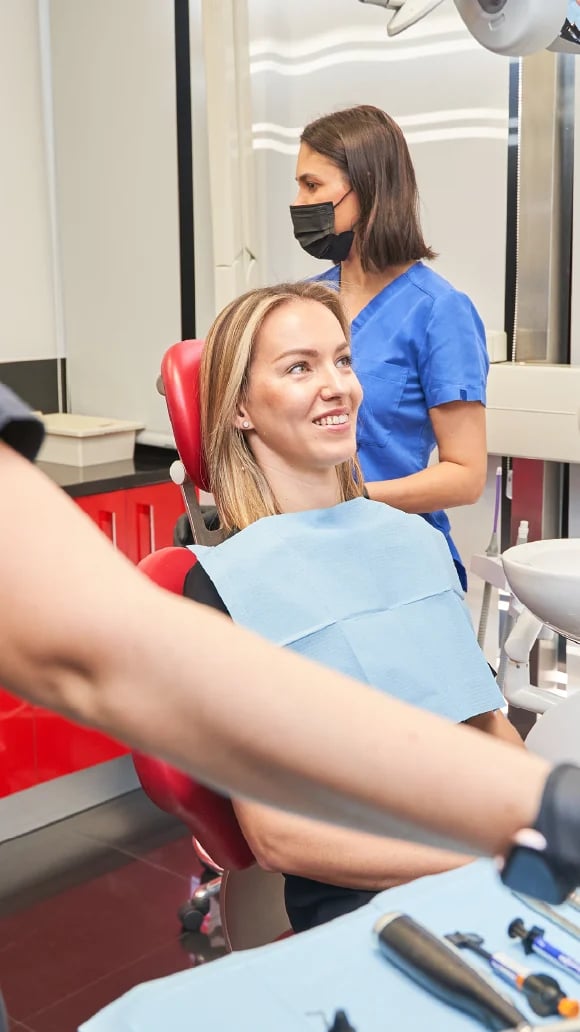Woman in dental chair smiling while dentist in blue scrubs and mask examines her teeth in modern dental office