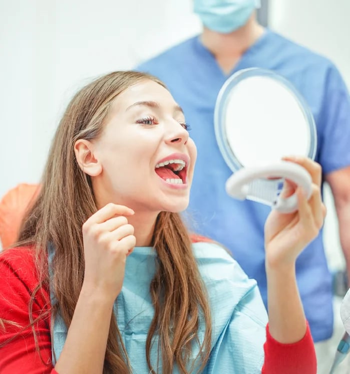 Patient woman admiring her smile in a mirror while a dentist in blue scrubs stands behind her in a dental office.