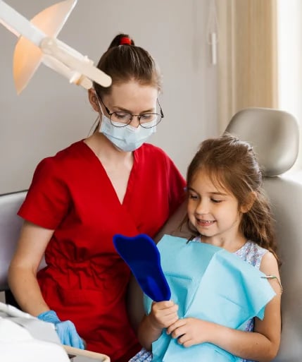 Pediatric dentist in red scrubs and mask examining a young girl patient in dental office chair